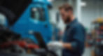 Mechanic in blue overalls using a laptop to inspect an engine in a garage. Blue truck in the background. Focused expression.