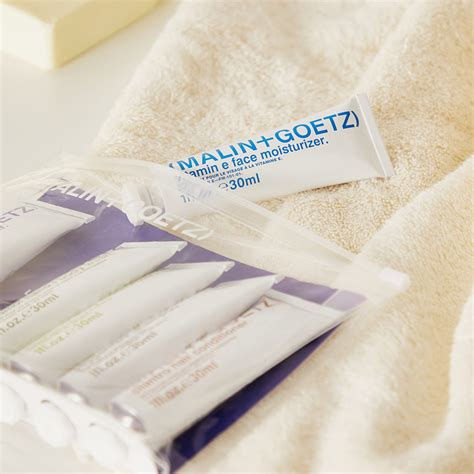 Toiletry bag and vitamin E face moisturizer tube on a soft beige towel. Text: MALIN+GOETZ. Calm, clean setting.