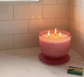 Pink candle with three flames on a marble surface beside a green matchbox. White tiled background creates a cozy ambiance.