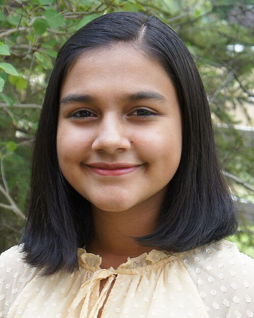 Smiling person with short black hair wearing a beige polka-dot blouse, standing outdoors with green foliage in the background. This is Gitanjali Rao.