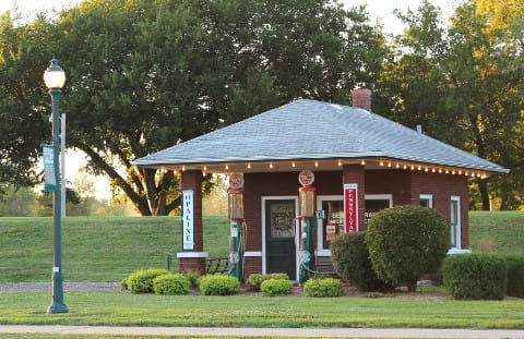 Historic Filling Station with brick exterior pillars and two historic filling pumps with shrubbery surrounding filing station with trees and grass in the background