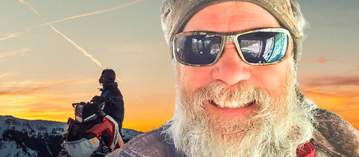 man taking a selfie with a man sitting on a snowmobile watches the sunset