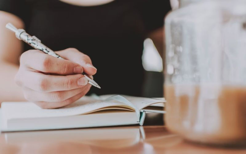 Hand writing in an open notebook beside a blurred coffee cup on a table, warm indoor study mood