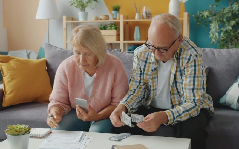 An elderly couple sits on a sofa reviewing finances. She holds a phone, and he counts money. The room has colorful cushions and shelves.
