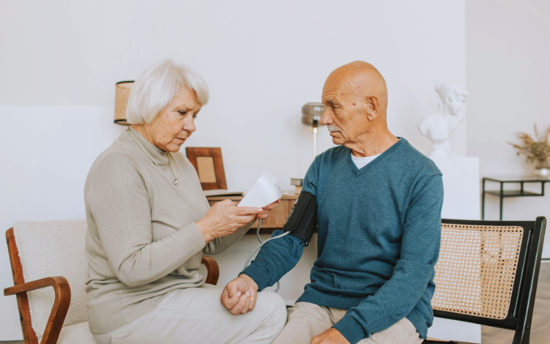 Elderly woman checks elderly man's blood pressure at home. He wears a blue sweater, she a beige one. Room is warmly lit, calm atmosphere.