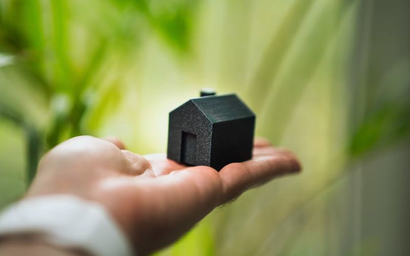 Hand holding a small black house model against a blurred green background, suggesting eco-friendly or housing concept.
