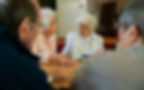 Elderly group conversing at a table in a cozy room. Women in white and peach engage attentively. Warm, intimate atmosphere.