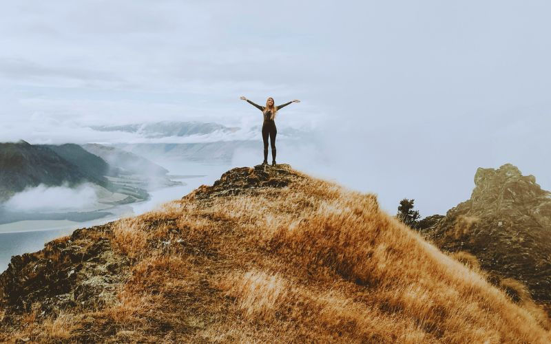 Person standing with arms outstretched on a grassy hilltop, overlooking a foggy landscape and distant mountains. Calm and triumphant mood.