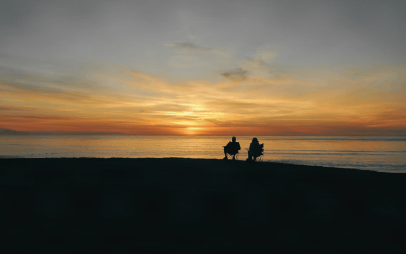 Two silhouetted people sit in chairs on a beach, watching a golden sunset over calm water.