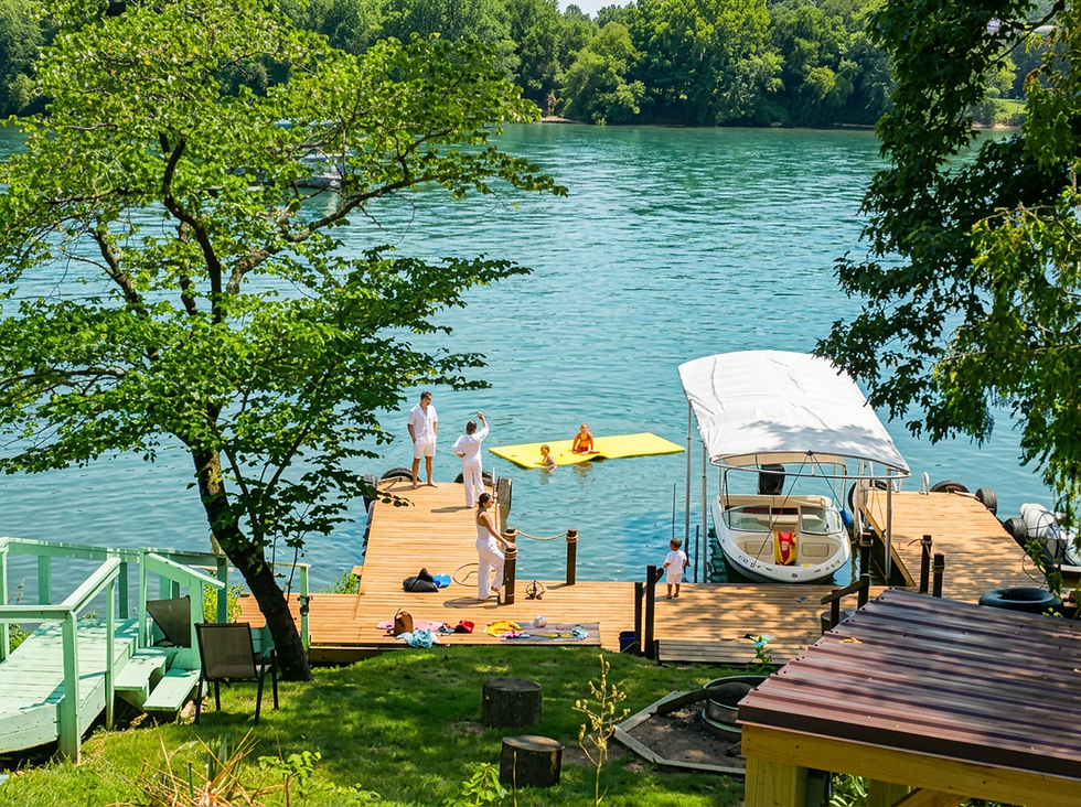 Outdoor photography, people relaxing on a wooden dock by a sunny lake. A peaceful summer atmosphere with a small boat moored nearby and lush trees surrounding the water.