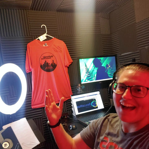 Brittany Tomes pictured excitedly in a selfie of her new and improved VO booth. Featuring red LED lighting, a ring light for self tapes and classes, acoustic foam paneling, lights, dual monitors, a Rode NT1-A microphone, Scarlet Interface, a standing desk, and a Bethesda Game Days Tshirt in the background. Brittany wears a NerdsVote tshirt and glasses, her blonde hair is pulled back in a ponytail.