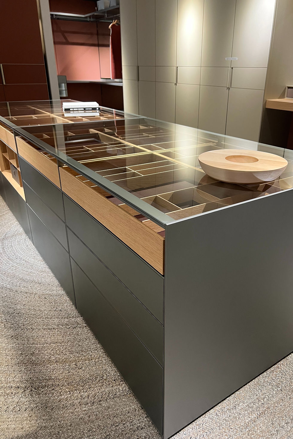 Modern showroom with a sleek glass-top display counter. Wooden bowl and books on top. Neutral-toned cabinets and carpeted floor. wardrobe island