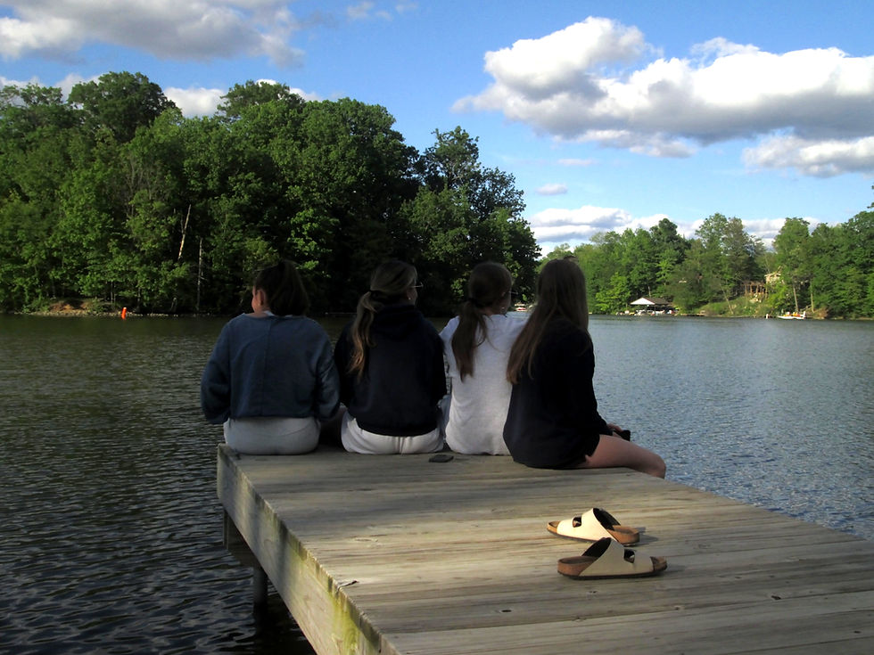 Four people sit on a wooden dock, gazing at a serene lake with lush trees and fluffy clouds. Sandals rest on the dock beside them.