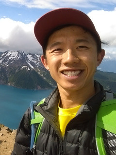 A man with a red baseball cap smiles at the camera. A lake and mountain range is visible in the background.