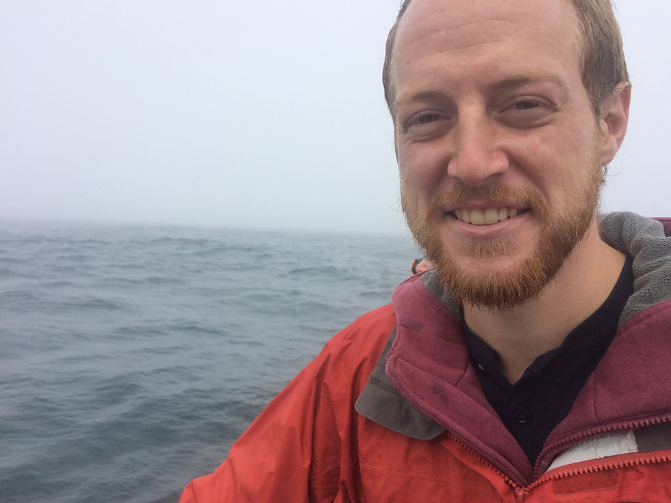 A bearded man smiles at the camera. There is water in the background (man appears to be on a boat). It is overcast.