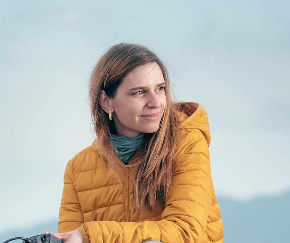 A woman with long hair smiles and looks off camera to right. She is sitting outside with a yellow puffer jacket holding a scientific water sampling instrument off camera