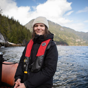 A woman with brown hair and a knit toque wears a black jacket and a life jacket and smiles at the camera. She is sitting in the bow of a zodiac on the water with steep tree covered hills and shorelines, typical of the Pacific northwest, in the background.