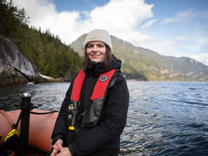 A woman with brown hair and a knit toque wears a black jacket and a life jacket and smiles at the camera. She is sitting in the bow of a zodiac on the water with steep tree covered hills and shorelines, typical of the Pacific northwest, in the background.
