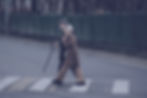 Elderly couple in coats walking hand in hand across a street crosswalk. Man uses a cane. Background shows green fence and bare trees.