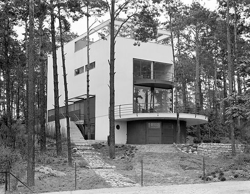 White building in a forest setting. There are large windows, a roof garden and semi-circular balconies on the front side.