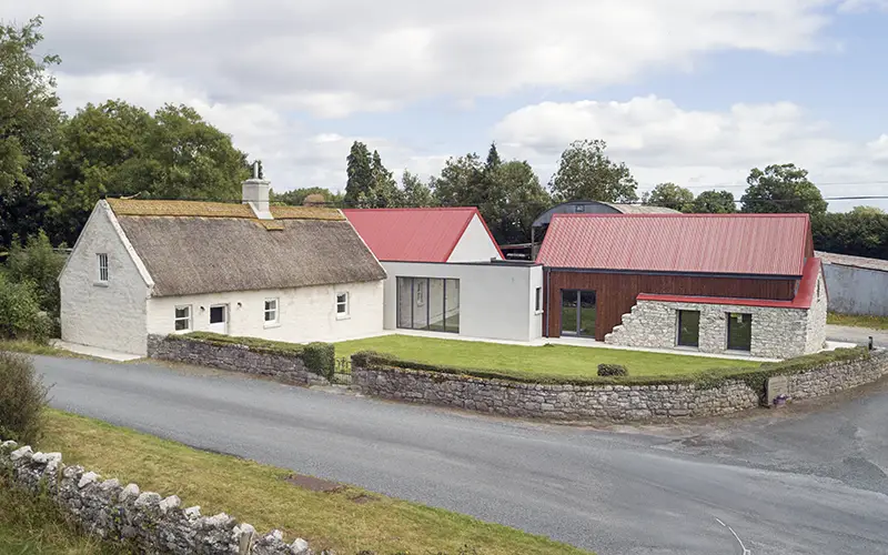 Traditional thatched cottage with a thatched roof next to a modern house with red roofs and large windows, set in a rural landscape.