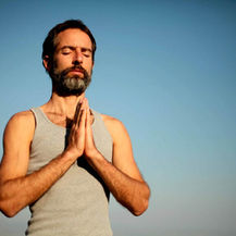Man in a gray tank top practicing meditation with hands in prayer position under a clear blue sky, symbolizing calm, focus, and mental resilience.