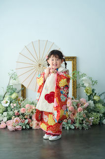 赤やオレンジの華やかな着物を着た3歳の女の子が、和傘を持って笑顔で立っている七五三写真。背景にはピンクや白のお花やグリーン、金色の額縁が飾られている。