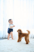 カーテンの前で、白いトップスと紺色のパンツを着た男の子が立ち、隣には茶色のトイプードルが立っている写真。