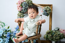 淡いグリーンの衣装を着た男の子が木製の椅子にスァ離、ブルーやグリーン、ピンクの紫陽花とアンティーク調の額縁に囲まれている