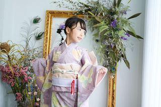 淡い紫色の花柄の着物を着た7歳の女の子が片方の袖を広げながら横を向いている七五三写真。背景には金色の額縁と、ピンクや紫のお花、グリーンが飾られている。