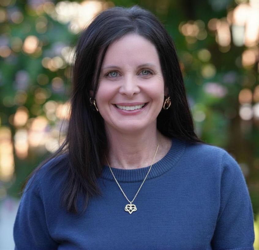 Smiling woman with long dark hair wearing a blue sweater and heart-shaped necklace, photog