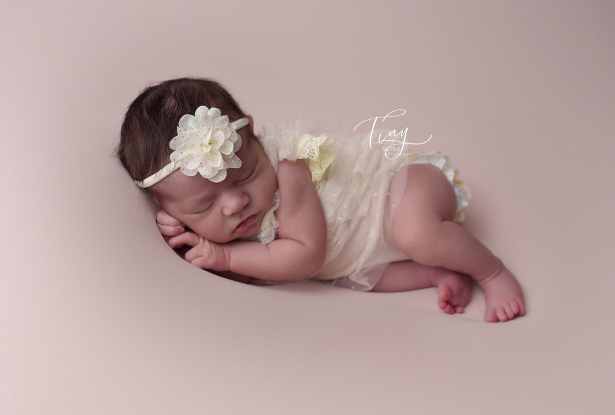 Newborn Baby Girl wearing a pretty cream dress and matching headband , sleeping in a side pose during the newborn photoshoot in Northern VA