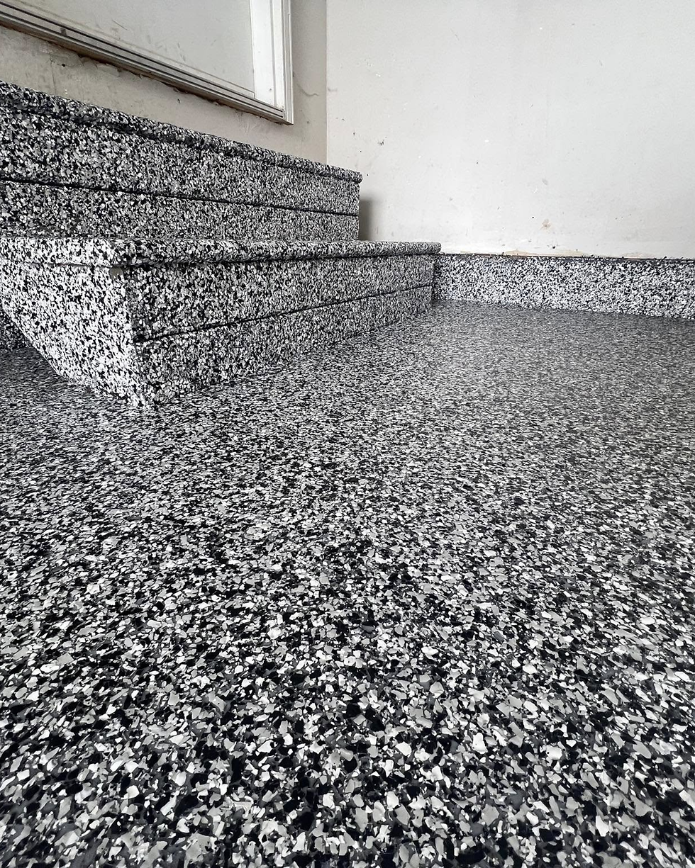 A side-by-side action shot of an installer wearing black gloves broadcasting decorative blue, gray, and white color chips to full rejection into a wet gray epoxy base coat.