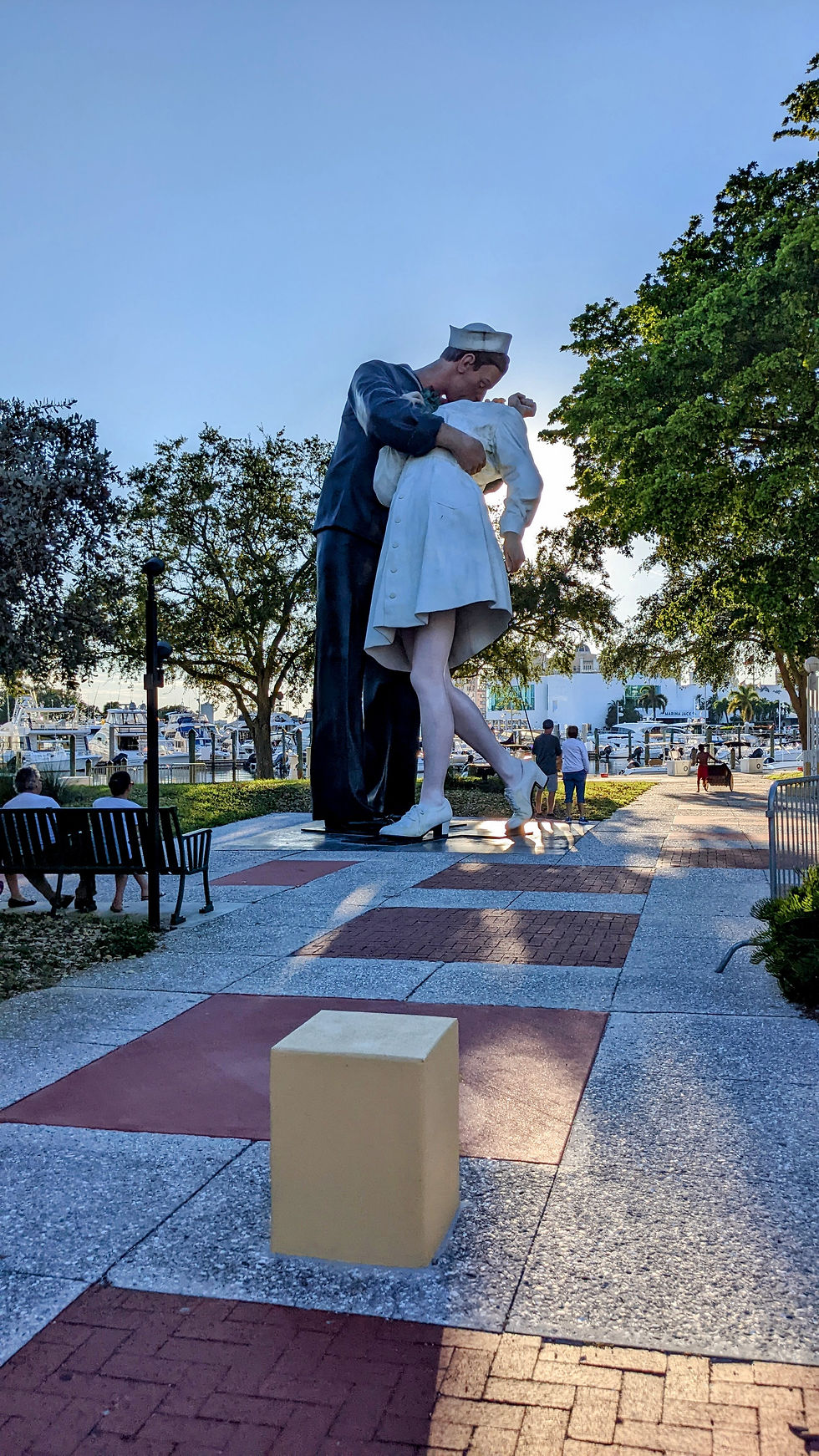 Large statue of a sailor kissing a nurse in a park with trees and benches. People walking dogs nearby. Blue sky background, sunny day.