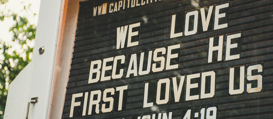 Church sign with white letters reads: "WE LOVE BECAUSE HE FIRST LOVED US 1 JOHN 4:19." Background is green foliage. Mood is inspirational.