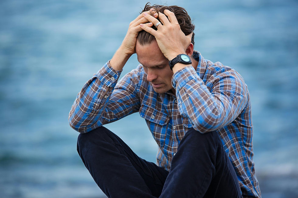 Man in plaid shirt sits by water with head in hands, appearing stressed. Background is a blurred blue expanse, mood is contemplative.