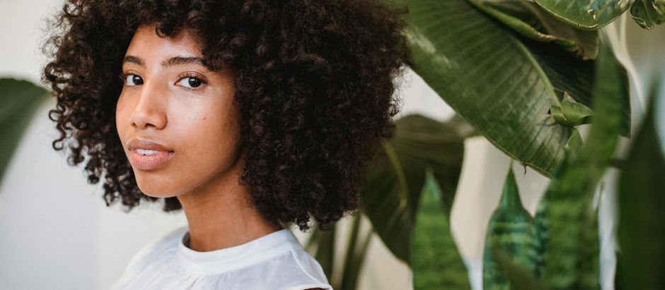 Woman with curly hair, wearing a white top, stands near large green plants. She gazes calmly at the camera, set against a bright background.