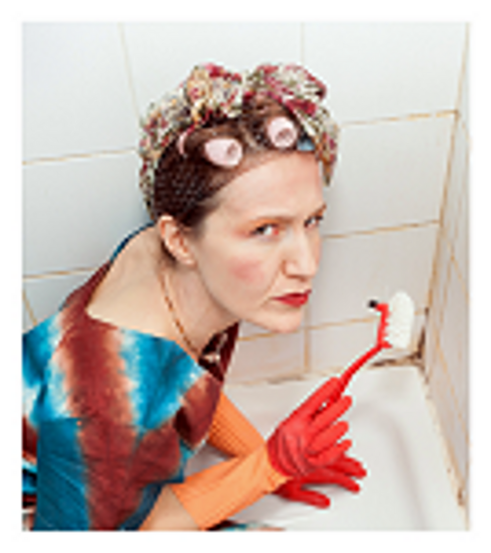 Mujer con rulos y guantes de goma limpia una bañera con cepillo. Viste vestido de colores. Mira determinada en un baño de azulejos blancos.
