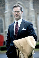 Business man in a natty dark blue suit carrying a camel trench coat in one arm.