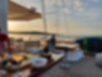 Wine glasses and dishes on a yacht table at sunset. A bottle in an ice bucket, paddleboard nearby. Calm sea and distant hills set the scene.