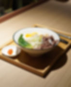 a wooden tray on a wooden table with a small bowl of chilli paste and a bowl of noodles with beef slices, leafy vegetables and hard-boiled eggs
