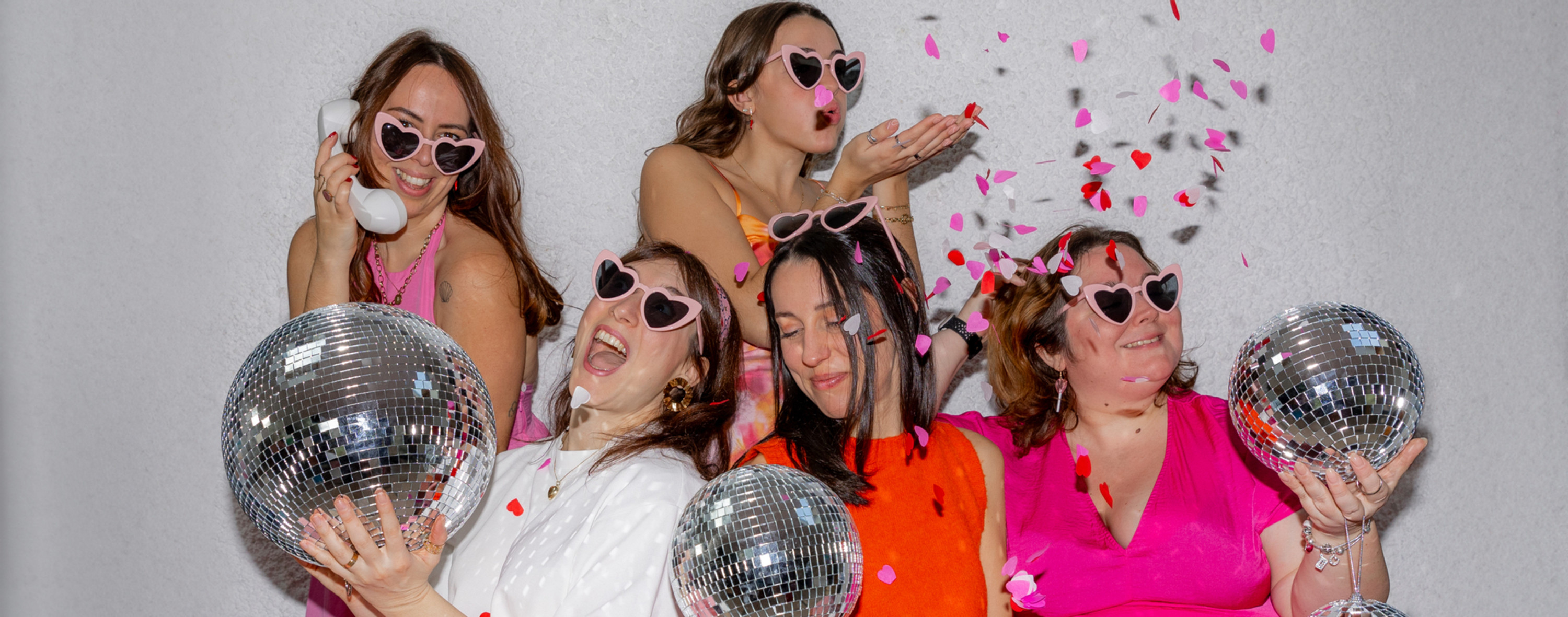 Cinq femmes wedding planner & designer souriantes en lunettes cœur posent avec des boules à facettes; confettis roses et rouges volent sur fond gris.