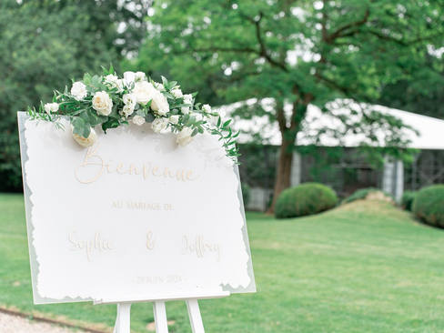 un tableau de bienvenue d'un mariage, il est blanc avec une écriture dorée et une composition florale posée dessus