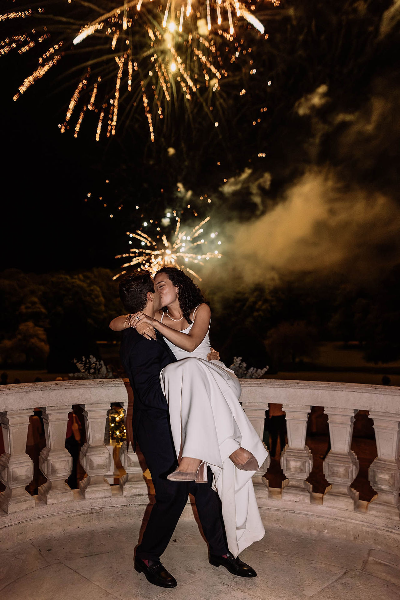 Deux mariés qui s'embrassent, la mariée dans les bras du marié avec un feu d'artifice en fond