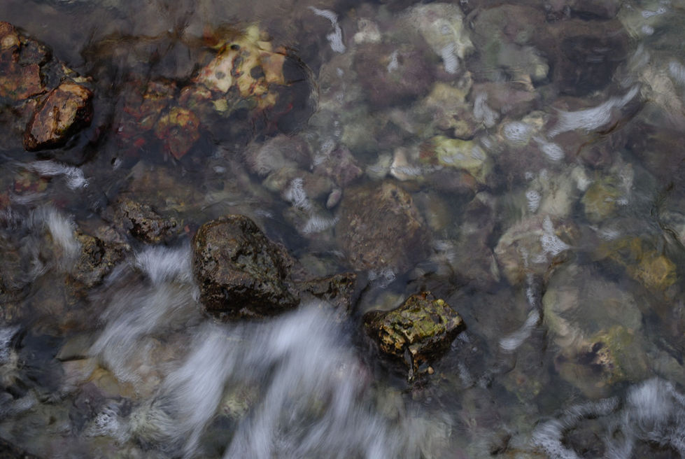 Clear water flows over smooth stones and rocks in a stream, creating gentle ripples and a peaceful, natural atmosphere.