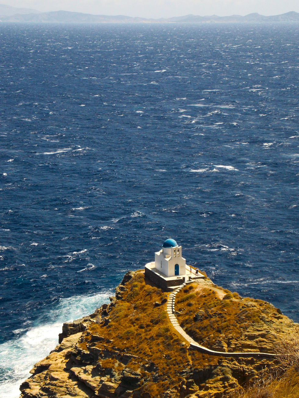 A small white chapel with a blue dome sits atop a rocky hill overlooking the vast, deep blue sea. A winding path leads to the chapel, set against a sunlit, rugged landscape.