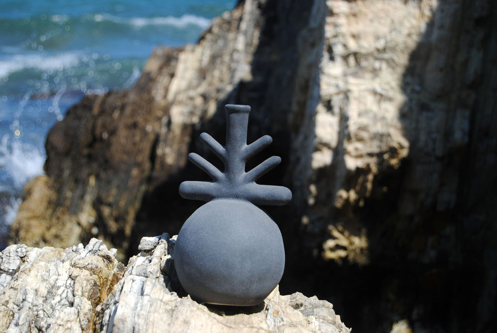 A uniquely shaped black sculpture resembling a bottle with protrusions rests on rocky seashore, with ocean waves in the background under a clear blue sky.