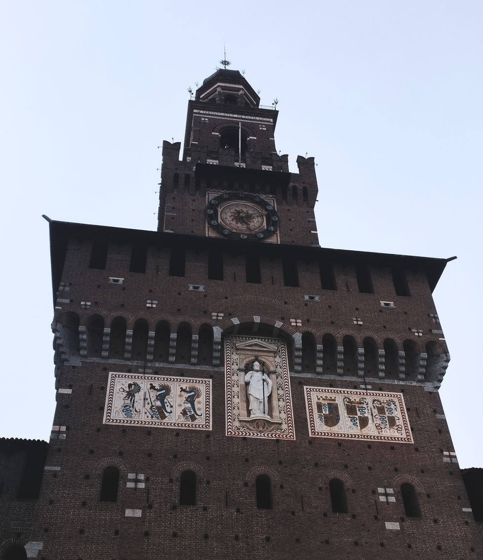 A towering brick castle facade featuring ornate frescoes and a central clock. The architecture conveys a historical and majestic atmosphere.