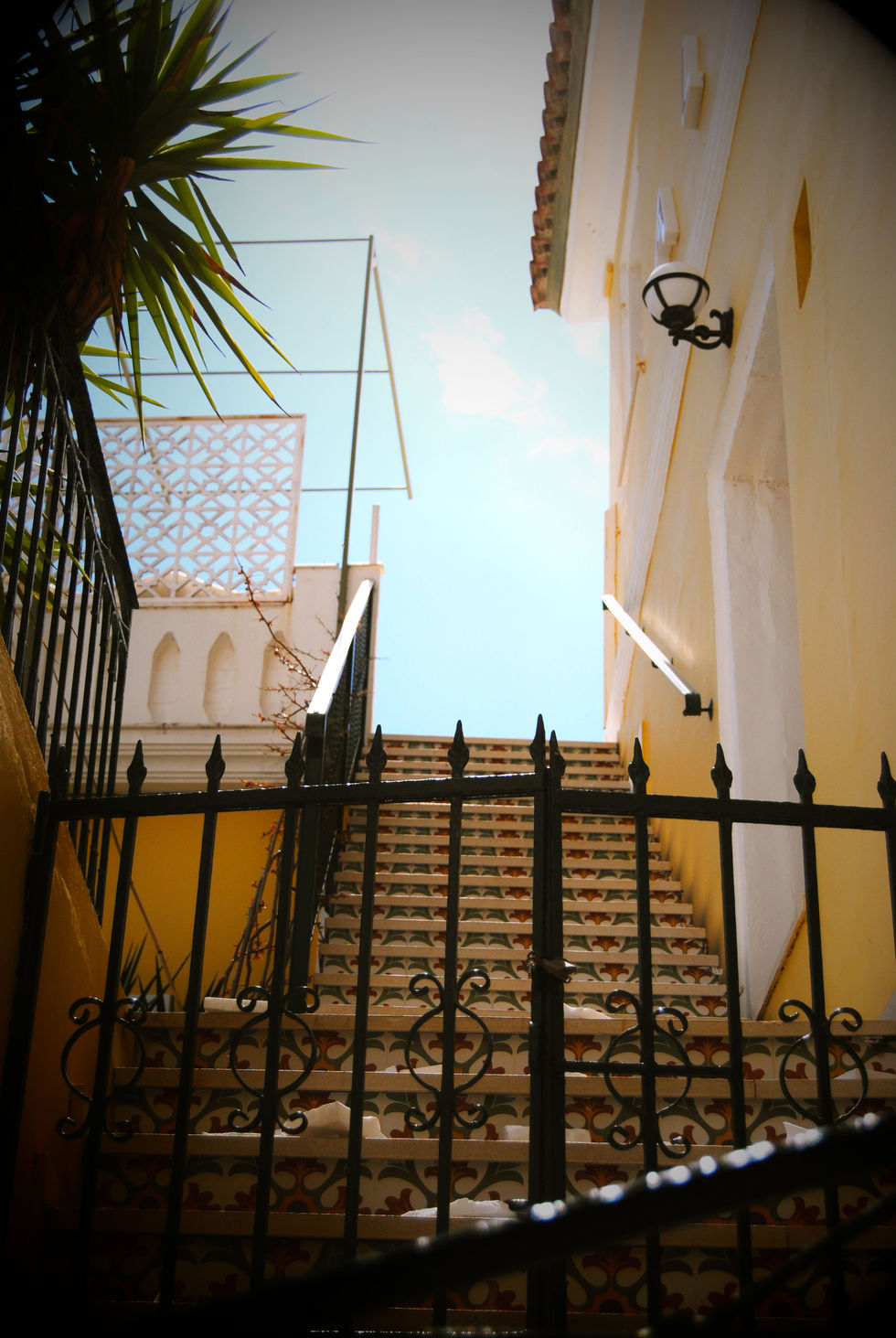 View of a bright, sunny courtyard with yellow walls, ornate black ironwork, palm fronds, and a clear blue sky, creating a tropical feel.