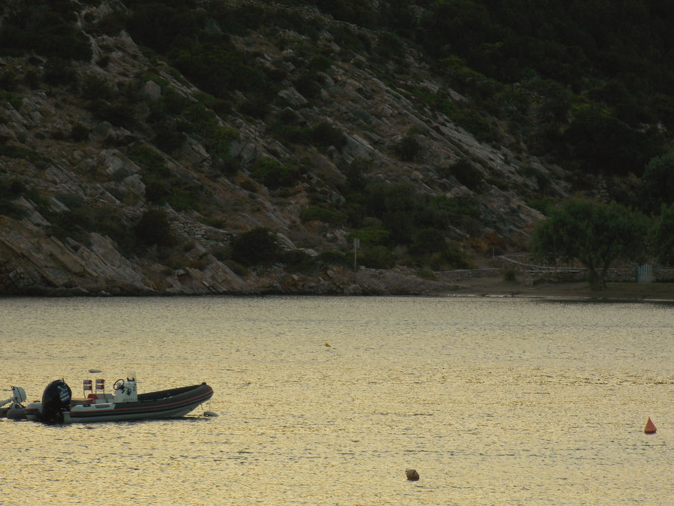 A small boat floats on calm, golden water at sunset, near a rocky shoreline with green vegetation. The scene is tranquil and serene.